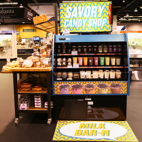 A standalone refrigerated display case filled with various prepared drinks and snacks. The display case has custom signs and matching floor decal.